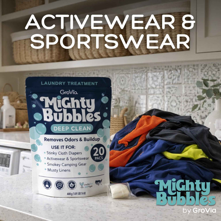 Mighty Bubbles laundry treatment packaging with a pile of colorful clothes on a kitchen counter.